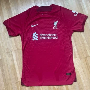 Liverpool FC röd Nike matchtröja - Liverpool FCs officiella matchtröja från Nike i en djup röd färg med subtilt geometriskt mönster. Tröjan har korta ärmar, rund hals och tryckta loggor från Standard Chartered och Expedia. Materialet är lätt och andas, perfekt för fotboll eller häng med polarna.
