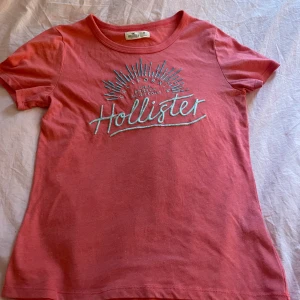 Rosa t-shirt från Hollister med brodyr - Snygg rosa t-shirt från Hollister med broderad text och solstråle-design på bröstet. Klassisk rund halsringning och korta ärmar. Perfekt för dig som gillar en avslappnad och cool stil. Tillverkad i mjuk bomull för skön känsla hela dagen.