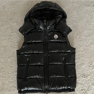 Svart dunväst från Moncler med huva - Svart dunväst från Moncler med glansig finish och huva. Västen har två dragkedjefickor, quiltad design och Moncler-logga på bröstet. Stängs med dragkedja framtill och har snygga detaljer som ger en exklusiv vibe. Perfekt för att lyfta din streetstyle.