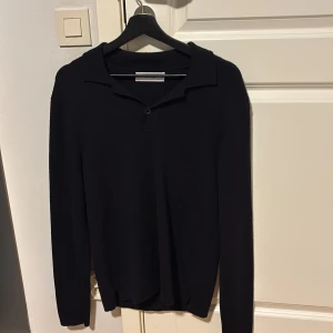 Svart stickad pikétröja Whyred - Säljer en svart stickad pikétröja från Whyred med klassisk krage och knapp framtill. Tröjan har lång ärm och är i mjukt material som passar perfekt till lager-på-lager under höst och vinter.  Merino ull