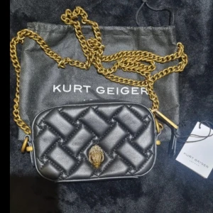 Svart quiltad axelväska Kurt Geiger - Snygg svart quiltad axelväska från Kurt Geiger med guldfärgad kedja och ikonisk örndetalj framtill. Väskan är i skinn och har en kompakt, rektangulär form som passar perfekt för det viktigaste. Kommer med dustbag och tagg.