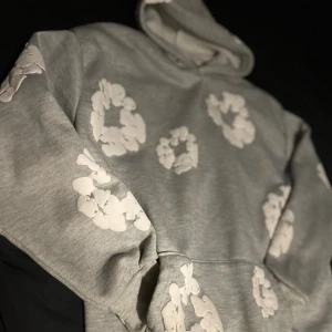 Grå hoodie från Denim Tears med blommor - Säljer en grå hoodie från Denim Tears med vita blommiga tryck över hela tröjan. Tröjan har huva och en klassisk känguruficka framtill. Dene rkt najs nu t hösten o vintern Storlek S men ja skulle säga att den sitter mer som M/L