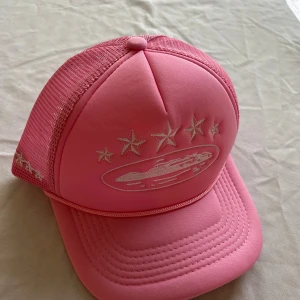 Rosa truckerkeps  Corteiz - Säljer en rosa truckerkeps  Corteiz med vit broderad logga och stjärnor framtill. Kepsen har mesh på sidorna och bak, samt böjd skärm. Insidan har band med Corteiz-tryck. Perfekt för dig som vill sticka ut med en färgstark accessoar.