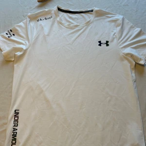 Vit Under Armour t-shirt för träning - Snygg vit t-shirt  Under Armour med svart logga på bröstet och tryck på ärmen. Tillverkad i lätt och ventilerande polyester, perfekt för löpning eller gym. Klassisk passform med rund hals och korta ärmar. Sportig och stilren design.
