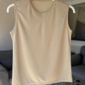 Beige ärmlös topp - En stilren beige topp med rund halsringning och ärmlös design. Toppen är tillverkad i ett mjukt och stretchigt material som ger en clean och enkel look. Perfekt att matcha med jeans eller kjol för en avslappnad vibe. Mindre fläck som skapar kontrast i färgen vid armhåla.