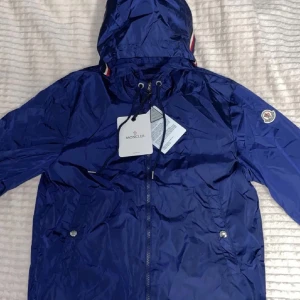 Blå vindjacka från Moncler med huva - Snygg blå vindjacka från Moncler med huva och dragkedja framtill. Jackan har Moncler-logga på ärmen, snörning vid huvan och diskreta fickor med tryckknappar. Materialet är glansigt och lätt, perfekt för blåsiga dagar.