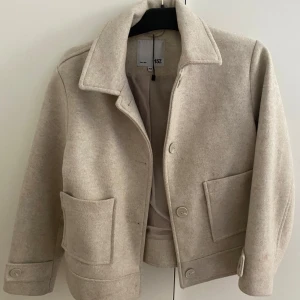 Beige kappa  - Stilren beige kappa  från 157 i ullmix med klassisk krage, stora fickor framtill och knäppning med knappar. Jackan har en rak passform och snygga detaljer vid ärmsluten. Perfekt för dig som gillar o vara lite extra och clean stil. Köpte i somras använd 3ggr. De står 160 men jag som har S-m kan ha den! Älskar 