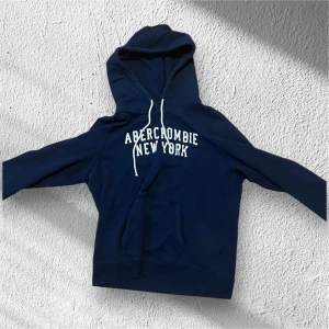 Marinblå hoodie Abercrombie & Fitch - Snygg marinblå hoodie från Abercrombie & Fitch med vit text 'Abercrombie New York' på bröstet. Tröjan har huva med vita snören och en stor magficka framtill. Perfekt för chill dagar och streetstyle.