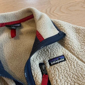 Beige fleecejacka från Patagonia - Säljer en beige fleecejacka från Patagonia med marinblå detaljer och röda dragkedjor. Jackan har hög krage, hel dragkedja framtill och tre fickor med dragkedja. Perfekt för kalla dagar och chill stunder utomhus.