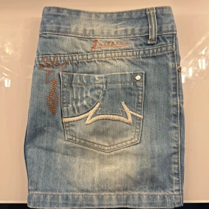 Blå jeanskjol från Just Play - Snygg ljusblå jeanskjol från Just Play med broderade detaljer och coola fickor bak. Kjolen har gul brodyr i midjan och orange broderi vid fickan. Klassisk femficksmodell med dragkedja och knapp framtill. Perfekt för dig som gillar streetstyle.💕💕💕