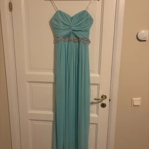 Ljusblå långklänning City Triangles - Säljer en ljusblå långklänning från City Triangles utan axelband och vackert draperad överdel. Klänningen har en glittrig dekoration i midjan och är tillverkad i mjuk polyester. Perfekt för bal eller student! Snyggt fall och romantisk känsla.