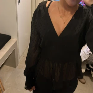 Svart blus med v-ringning och volang - Säljer en svart blus med v-ringning och volang nedtill. Blusen har långärmade, lätt transparenta ärmar med diskret prickigt mönster och knappar vid ärmsluten. Sitter snyggt i midjan och har en luftig, flowy känsla.