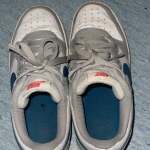Nike Air Force 1 vita och blå sneakers - Säljer ett par klassiska Nike Air Force 1 sneakers i vitt och blått med grå detaljer. Skorna har röd Nike-logga på plösen, snörning och perforerad tå för extra ventilation. Perfekta för dig som gillar streetstyle och vill ha en ikonisk look. Sizen är 38 EU