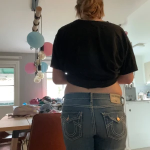 True Religion blå bootcut jeans - Säljer ett par blå jeans från True Religion med ikoniska bakfickor och kontrastsömmar. Modellen har låg midja och bootcut-ben, klassisk denim med snygga slitningar och detaljerade fickor. Perfekta för dig som vill sticka ut med en streetig look.