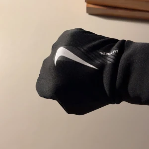 Nike svarta Therm-Fit träningshandskar - Svarta Nike Therm-Fit handskar med vit swoosh-logga på ovansidan. Insidan har greppvänliga prickar för bättre fäste. Smidig och sportig design med tight passform, perfekta för träning utomhus när det är kallt. Materialet är syntet och håller värmen bra.