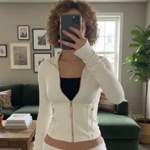 Vit vindjacka från Lululemon - Snygg vit vindjacka från Lululemon med guldiga dragkedjor och högt krage. Jackan har figurnära passform, långa ärmar och två fickor med dragkedja. Perfekt för träning eller chill dagar. Lululemon-logga på ryggen och coola sömdetaljer framtill.