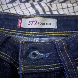 Levi's 572 Bootcut jeans mörkblå - Säljer ett par Levi's 572 Bootcut jeans i mörkblå tvätt. Klassisk femficksmodell med kontrastsömmar och bootcut-ben som ger en snygg siluett. Jeansen har Levi's patch bak och röd etikett vid fickan. Perfekt för dig som gillar retrovibbar och tidlös denim.