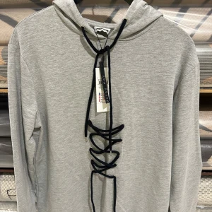 Grå hoodie med snörning från Noisy May - Säljer en grå hoodie från Noisy May med svart snörning framtill som cool detalj. Tröjan har huva och långa ärmar, perfekt för en avslappnad och trendig look. Materialet är en mjuk mix av bomull och polyester.