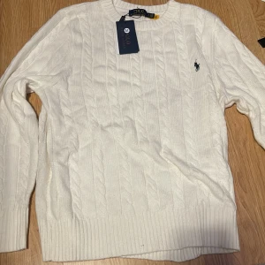  Polo Ralph Lauren - Kabelstickad vit tröja från Polo Ralph Lauren med klassisk rund hals och broderad marinblå logga på bröstet. Tröjan har ribbade muddar och är tillverkad i mjuk bomull. Perfekt för en clean och stilren look.