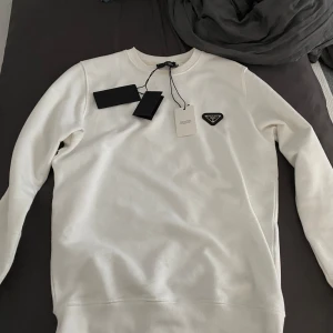 Vit tröja från Prada - Säljer en stilren vit tröja från Prada. Tröjan har en klassisk design med långärmade ärmar och en diskret Prada-logga på bröstet. Perfekt för både vardag och festliga tillfällen. Passar bra till alla säsonger och ger en lyxig touch till din outfit.