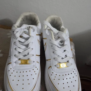 Nike CR7 vita och guld sneakers - Nike CR7 sneakers i vitt med snygga gulddetaljer och klassisk swoosh på sidan. Skorna har snörning, perforerad tå och CR7-logga bak. Tillverkade i syntetmaterial med vadderad insida för extra komfort. Perfekta för dig som gillar sportig och clean stil.