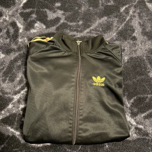 Svart Adidas track tjocktröja med guldiga ränder - Limited edition svart Adidas track jacket med guldiga ränder längs ärmarna och broderad Adidas-logga på bröstet samt stor logga på ryggen. Tröjan har dragkedja framtill, ribbade muddar och klassisk sportig look. Perfekt för dig som gillar streetwear och retrostil.