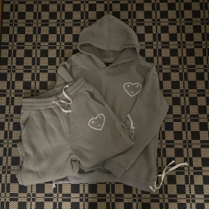 Grå hoodie och byxa med hjärtdetalj Carsicko - Grå hoodie från Carsicko med huva och lång ärm. På bröstet finns ett vitt hjärta med broderad smiley. Tröjan har ribbade muddar och dragsko nertill. Perfekt för chill dagar och snygg att matcha med mjukisbyxor.