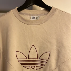 Beige sweatshirt från Adidas Originals - Snygg beige sweatshirt från Adidas Originals med det klassiska Trefoil-loggan broderad i vinrött på bröstet. Tröjan har rund hals, långa ärmar och ribbade muddar. Perfekt för chill dagar och streetwear-stil.