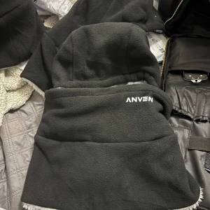 Svart fleece balaclava från Anven - Säljer en svart balaclava från Anven i mjuk fleece. Huvan täcker hela huvudet och halsen, med grå kant runt ansiktsöppningen. Perfekt för kalla vinterdagar och outdoor-aktiviteter. Snygg, enkel design med Anven-logga framtill.
