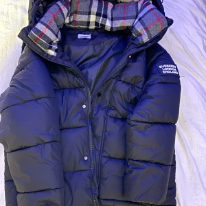 Svart pufferjacka Burberry M - Svart pufferjacka från Burberry London England med avtagbara ärmar och huva. Insidan har ett grått, vitt och rött rutigt foder. Jackan har dragkedja, tryckknappar och två fickor med dragkedja framtill. Perfekt för kalla dagar och riktigt snygg streetstil.