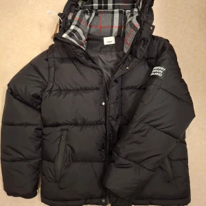 Svart pufferjacka från Burberry - Svart pufferjacka från Burberry med huva och dragkedja. Insidan har ett rutigt foder i grått, vitt och rött. Jackan har Burberry London England-logga på ärmen, två sidofickor med dragkedja och quiltad design. Perfekt för kalla dagar och riktigt snygg stil. S men passar M också