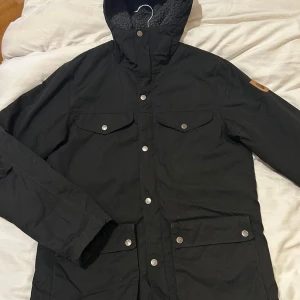 Svart parkajacka från Fjällräven - Svart parkajacka från Fjällräven med huva och mysigt teddyfoder. Jackan har fyra framfickor med tryckknappar, snygga metallknappar med logotyp och ett brunt lädermärke på ärmen. Perfekt för kalla dagar och har en stilren, lång design. Sparsamt använd 