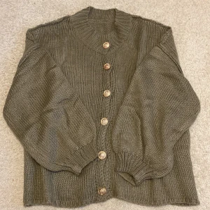 Olivgrön stickad kofta med guldknappar - Snygg olivgrön stickad kofta med  ribbad krage och sex dekorativa guldknappar framtill. Koftan har långärmade och breda muddar vid ärmslut och nederkant. Perfekt för att ge din outfit en chill och trendig vibe.