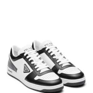 Svarta och vita sneakers från Prada - Snygga sneakers från Prada i svart och vitt skinn med klassisk låg siluett. Skorna har rund tå, snörning och perforerade detaljer på tån. Prada-logga på sidan och plös. Perfekt för dig som gillar exklusiv streetstyle.