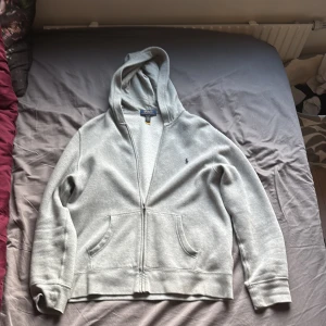 Grå hoodie från Polo Ralph Lauren - Snygg grå hoodie från Polo Ralph Lauren med klassisk broderad logga på bröstet. Tröjan har huva, dragkedja framtill och två stora fickor. Tillverkad i mjuk bomullsmix som är perfekt för chill dagar. Passformen är normal och ärmarna är långa.