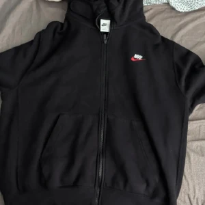 Svart Nike hoodie med dragkedja - Svart hoodie från Nike med klassisk logga i vitt och rött på bröstet. Tröjan har huva med snörning, hel dragkedja framtill och två stora fickor. Tillverkad i en mjuk blandning av bomull och polyester, perfekt för chill dagar eller när du vill ha en sportig look.