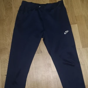 Mörkblå mjukisbyxor från Nike - Snygga mörkblå mjukisbyxor från Nike med klassisk logga på vänster ben. Byxorna har snörning i midjan och är tillverkade i mjukt material som är perfekt för chill eller träning. Passformen är normal och benen är raka med en modern look.