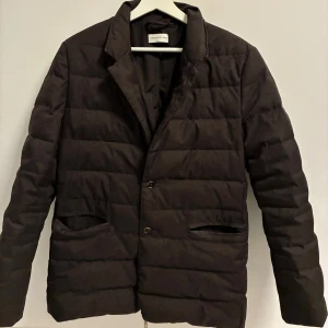 Brun quiltad kavajjacka These Glory Days - Stilren svart quiltad jacka från These Glory Days i kavajmodell. Jackan har två knappar framtill, klassisk krage och två fickor. Perfekt för dig som vill ha en dressad look men ändå hålla dig varm. Materialet är mjukt och känns lyxigt. Liknade Herno, nypris 3000kr