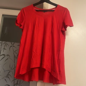 Röd tröja från Tommy Hilfiger  - Säljer en röd tröja från Tommy Hilfiger med rund halsringning och korta ärmar. Tröjan har en längre bakdel för en snygg siluett och är gjord i mjuk bomull. Perfekt för dig som gillar färgstarka och bekväma toppar med enkel design.