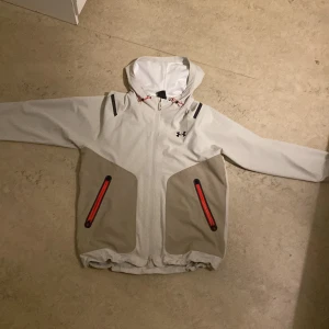 Under Armour vindjacka med huva - Ljusgrå och beige vindjacka från Under Armour med huva och röda detaljer vid fickorna. Jackan har hel dragkedja framtill, meshfoder i huvan och svarta loggan på bröstet. Perfekt för blåsiga dagar och sportiga outfits.