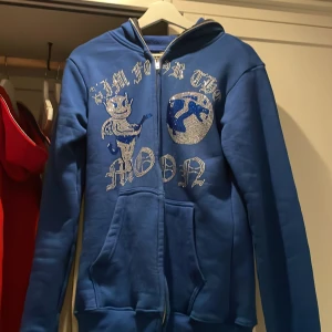 Blå hoodie Aim For The Moon M - Blå hoodie med dragkedja från Aim For The Moon. Framsidan har ett stort tryck med text och motiv i silver, samt fickor och huva. Mjuk insida och klassisk passform, perfekt för dig som gillar streetwear och vill sticka ut.