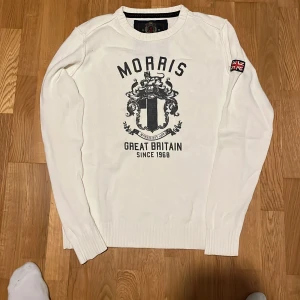 Vit stickad tröja Morris M - Snygg vit stickad tröja från Morris i storlek medium med svart tryck framtill och brittisk flagga på ärmen. Tröjan har rund hals och ribbade muddar vid ärmslut och nederkant. Perfekt för kallare dagar! Fint skick och storlek M, hör av dig vid några frågor!