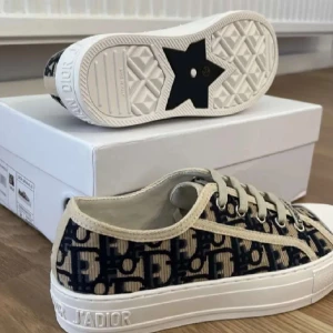 Dior sneakers med monogram och snörning - Snygga Dior sneakers med beige och marinblått monogrammönster, vita sulor och snörning. Skorna har tydlig Dior J'ADIOR-logga på sulan och är tillverkade i canvas med detaljer i bomull. Perfekta för dig som vill sticka ut med lyxig streetstyle.