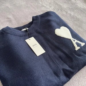 Ami Paris tröja  - Snygg marinblå stickad tröja från Ami med rund hals och ribbade muddar. På bröstet finns ett vitt hjärta med ett A, som är typiskt för märket. Perfekt för dig som gillar stilrena detaljer och vill ha något som sticker ut lite extra.