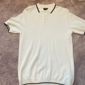 Vit piké från Oscar Jacobson, M - Snygg vit pikétröja från Oscar Jacobson i storlek M. Tröjan har klassisk krage med svarta detaljer, korta ärmar och tre knappar framtill. Materialet är mjukt och känns lyxigt mot huden. Perfekt för dig som gillar stilrena och enkla plagg. Tröjan är i mycket bra skick och har endast användits typ 1 gång