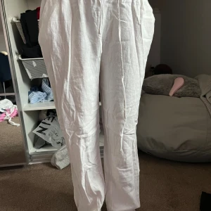 Vita linnebyxor med rak passform - Säljer ett par vita linnebyxor med avslappnad rak passform och resår i midjan. Byxorna är lätta och luftiga, perfekta för varmare dagar. De har en enkel och stilren look som funkar till många olika outfits.