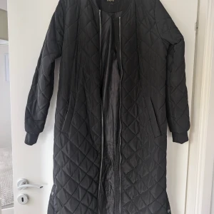 Svart quiltad lång jacka från ONLY - Lång svart quiltad jacka från ONLY med dragkedja framtill och tryckknappar i sidorna. Jackan har ribbade muddar vid halsen och ärmarna, samt två fickor. Perfekt för kyliga dagar och har en stilren look.