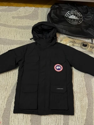 Canada Goose - Säljer en svart dunjacka från Canada Goose med huva och flera stora fickor framtill. Jackan har klassisk logotyp på bröstet och är fodrad med dun för extra värme. Perfekt för kalla dagar och har en stilren, tidlös design. Helt ny