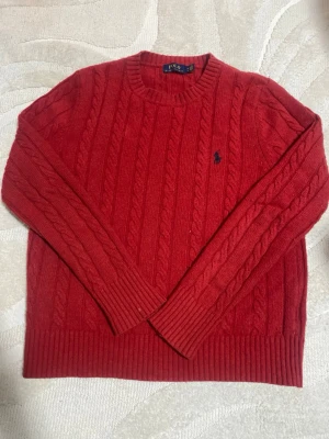 Polo Ralph Lauren Sweatshirt  - Kabelstickad röd tröja från Polo Ralph Lauren med rund halsringning och klassisk marinblå logga broderad på bröstet. Tröjan har långa ärmar och ribbade muddar vid ärmslut och nederkant. Perfekt för dig som gillar tidlös och snygg stil. Skickas samma dag, pris kan diskuteras 