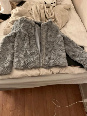 Ljusgrå pälsjacka/kappa - Säljer en ljusgrå pälsjacka/kappa med vågigt mönster och rund halsringning. Jackan har knäppning framtill och är fodrad på insidan. Perfekt för kalla dagar när du vill vara både varm och snygg.